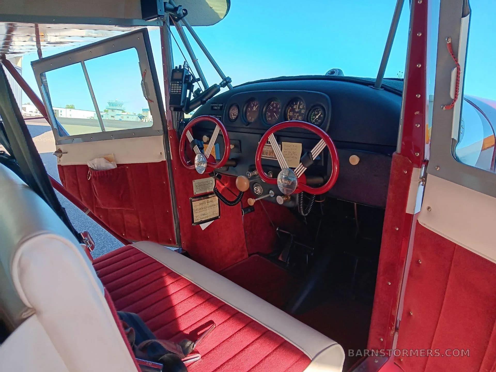The red and white interior cabin of an Aeronca Chief, showing the seats, instrument panel with red yokes, and an open door revealing an airport tower.