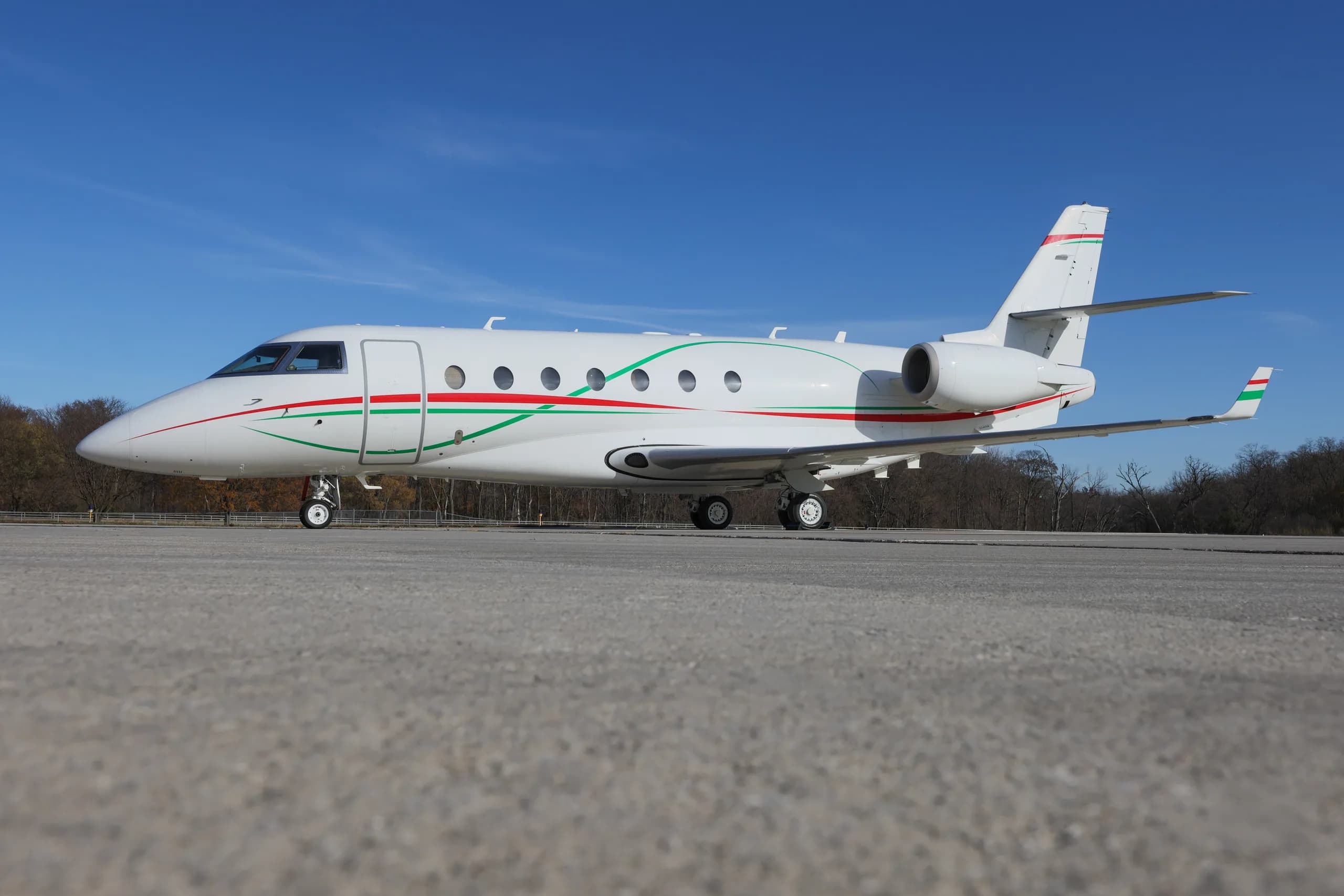 A white business jet with green and red stripes on its fuselage is parked on an asphalt runway under a clear blue sky.