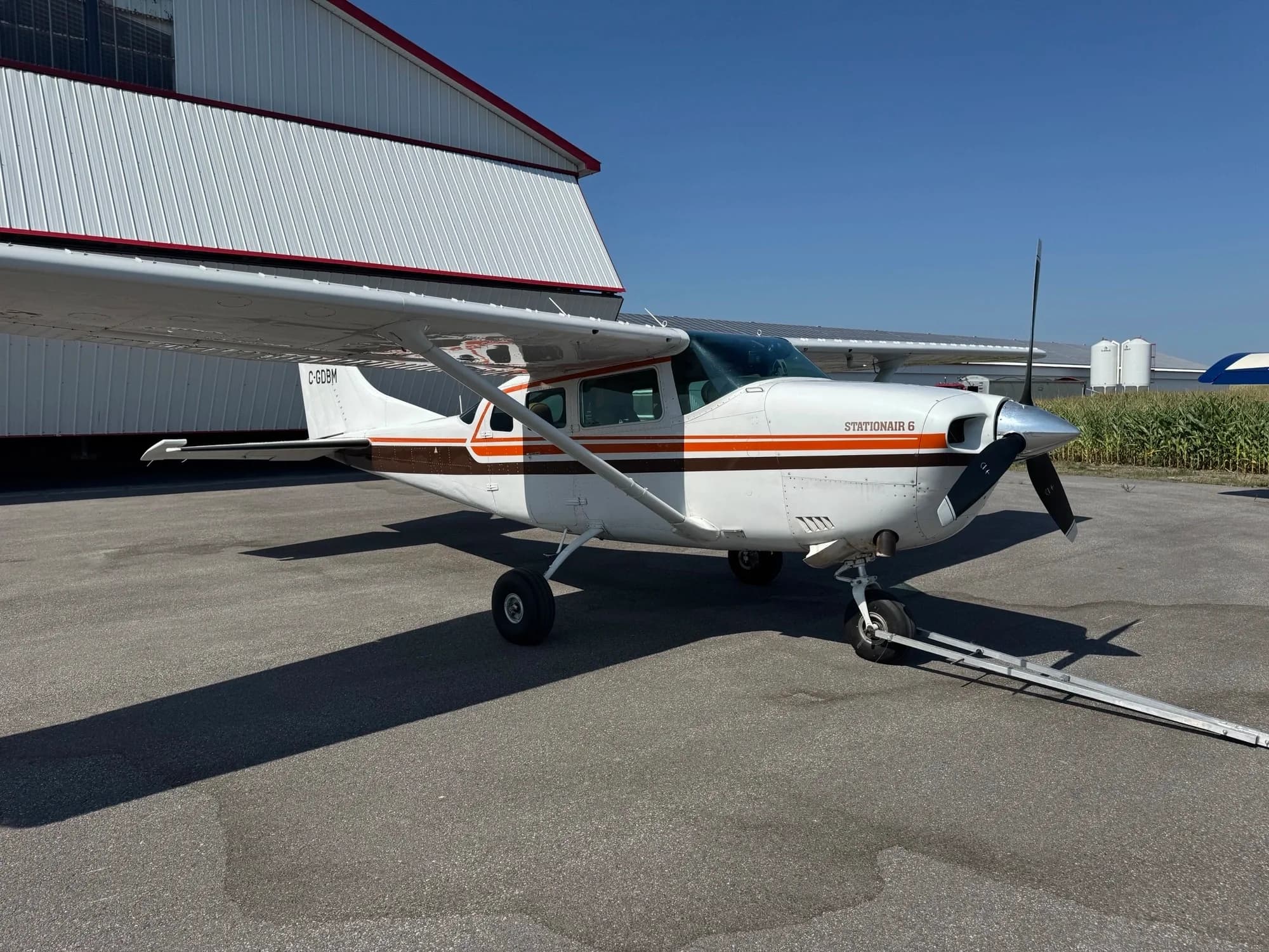 A white Cessna Stationair 6 with brown and orange stripes is parked on an asphalt ramp next to a white hangar under a clear blue sky.