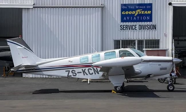 A white Beechcraft Baron with blue and red stripes is parked on a tarmac in front of a hangar with a Goodyear Aviation sign.