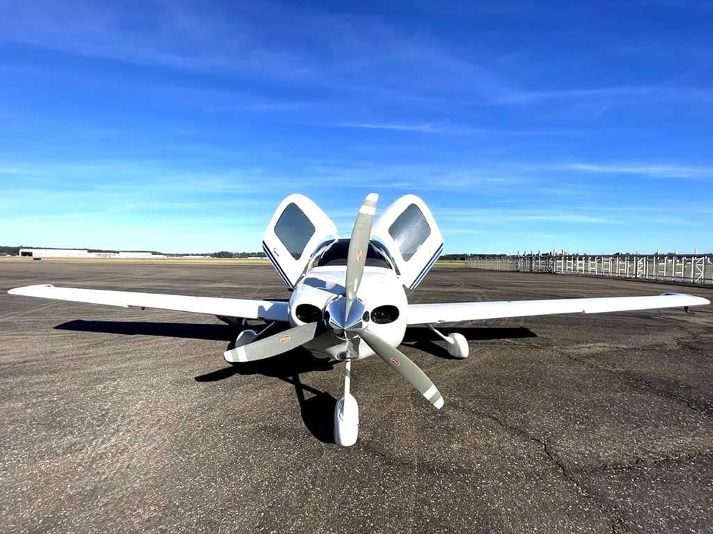 A head-on view of a white Cirrus SR20 aircraft, registration N824CD, with its doors open, parked on an asphalt runway under a clear blue sky.