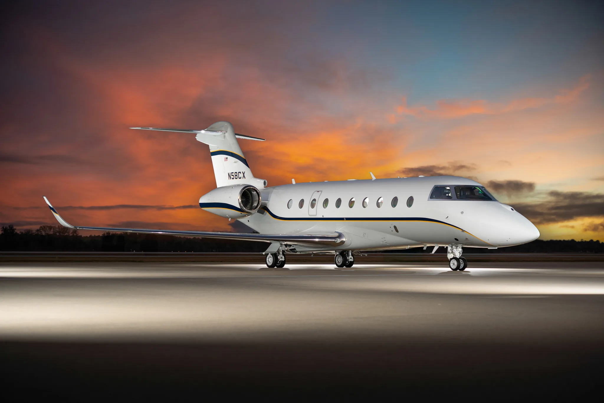 A white business jet with blue and gold stripes, registration N58CX, is parked on a tarmac at sunset with dramatic lighting.