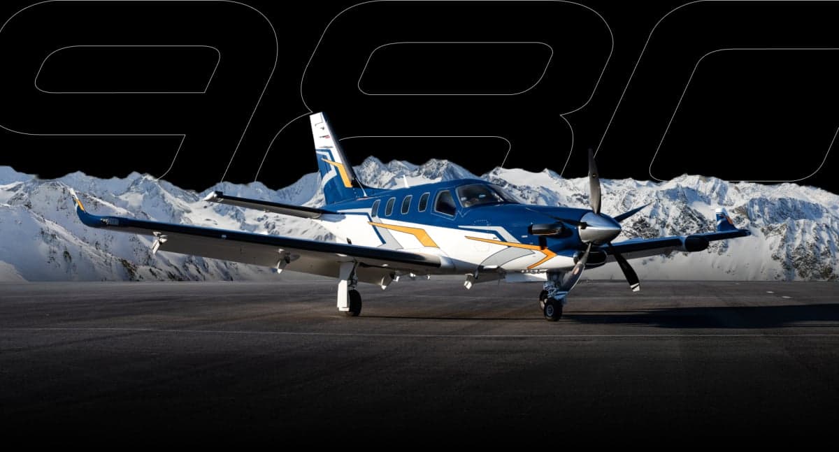A blue, white, and orange TBM 980 turboprop aircraft parked on an airfield with snowy mountains in the background.