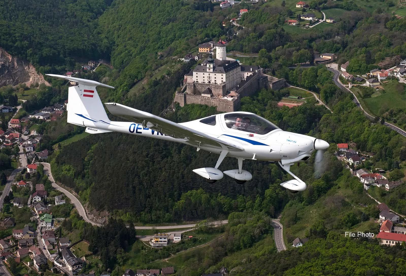 A white Diamond DA40 aircraft with blue stripes and registration OE-JYC flying over a green, hilly landscape with a town and a castle.