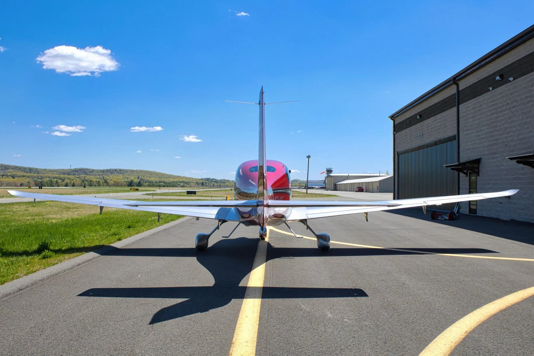 Rear view of a red and silver Cirrus SR22T aircraft parked on an airport tarmac with a hangar in the background.