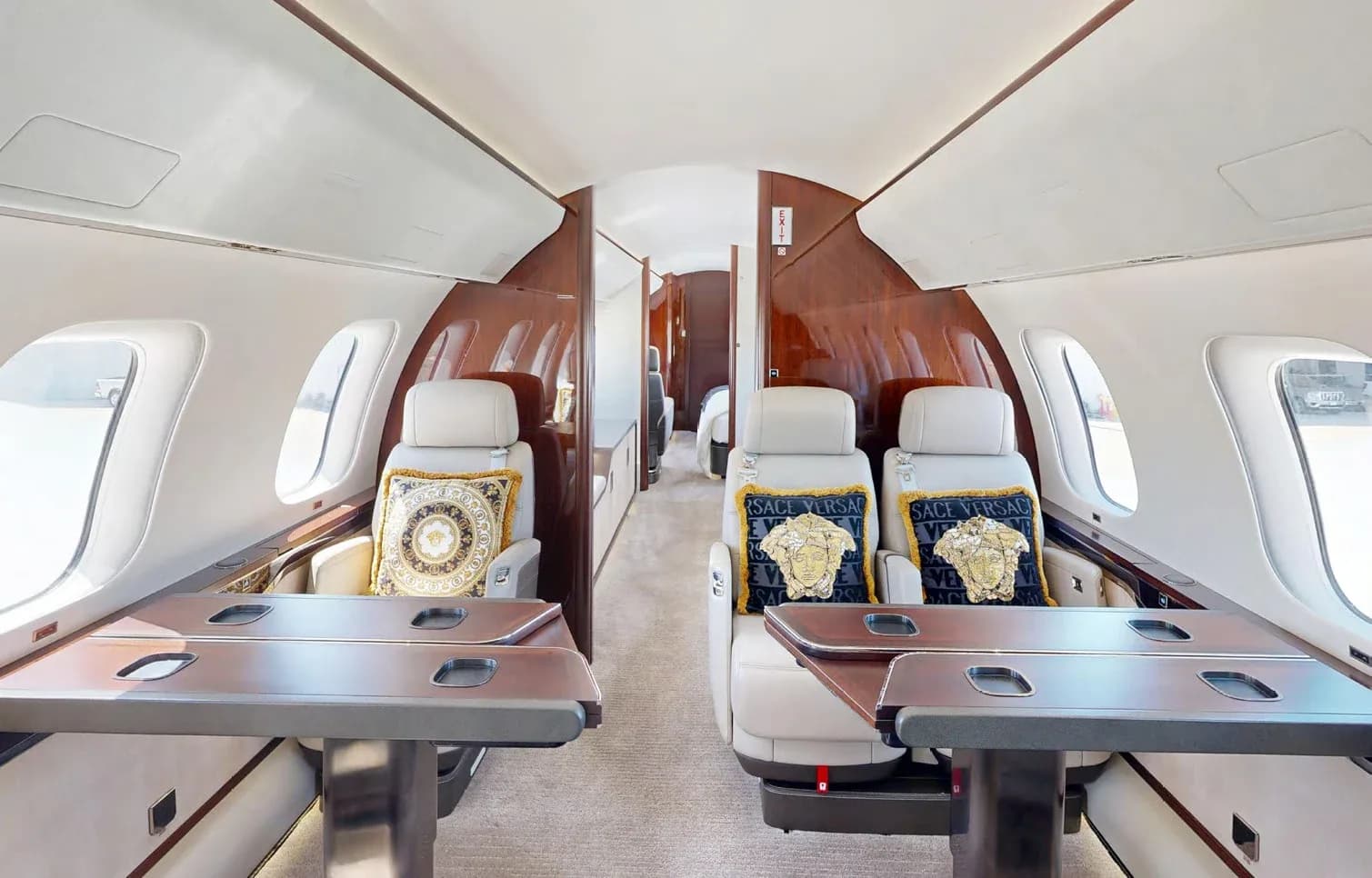A view inside the private jet cabin, showing two sets of facing seats with Versace pillows and polished dark wood tables.
