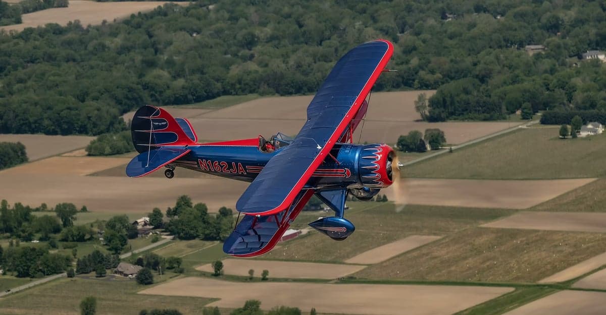 A red and blue biplane flies over a rural landscape.