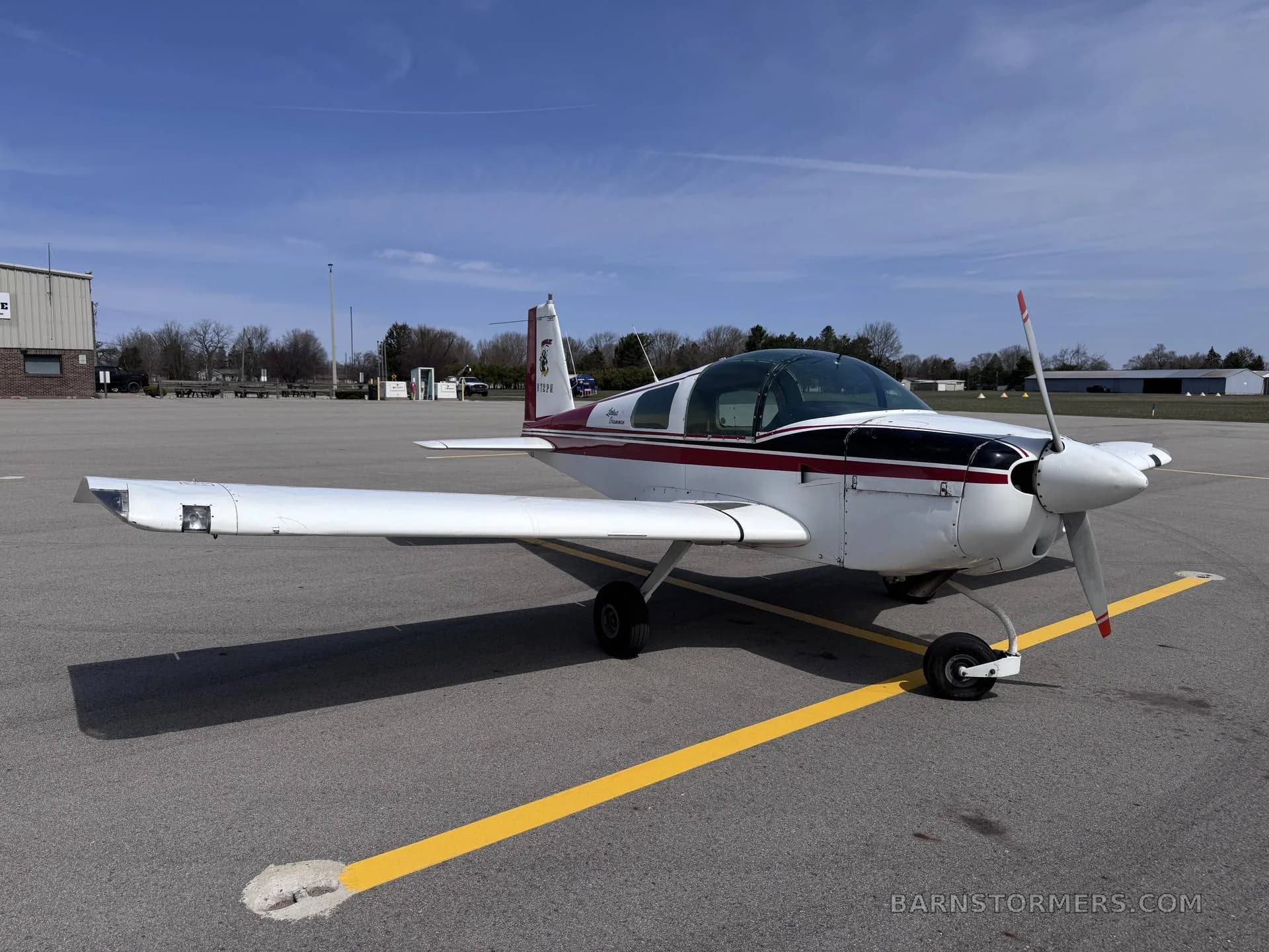 A white and maroon Grumman American AA-5 Traveler aircraft is parked on an asphalt tarmac under a clear blue sky.