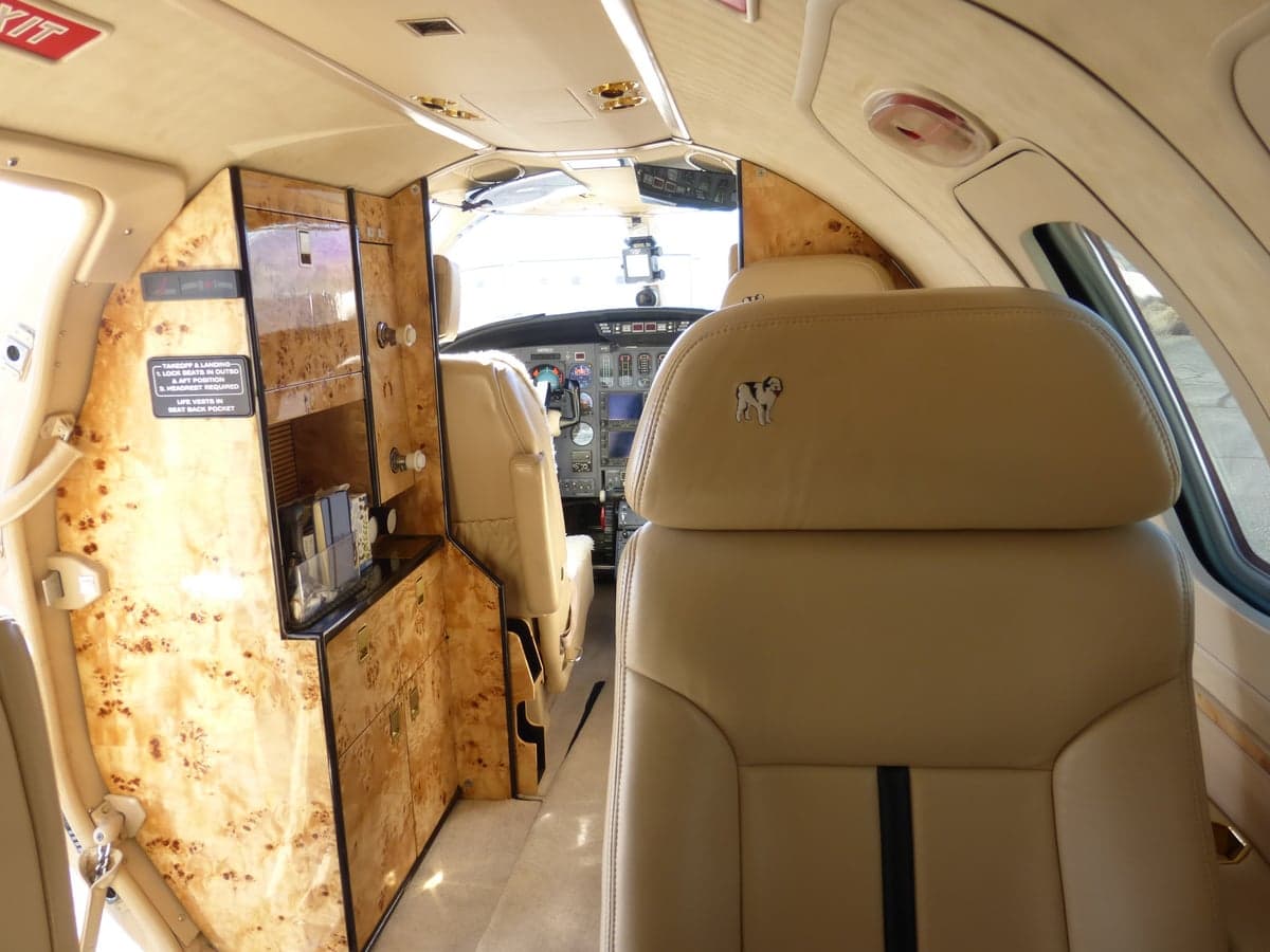 The luxurious interior cabin of a private jet, with tan leather seats, burled wood cabinetry, and a view towards the cockpit.