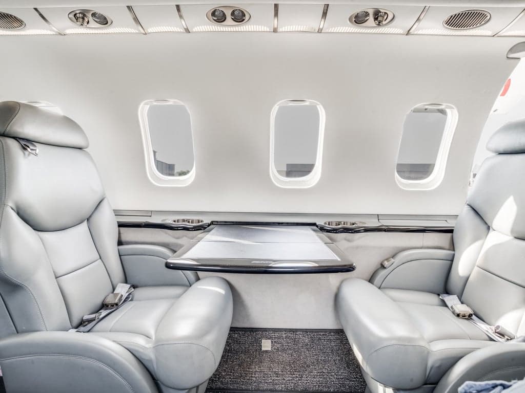 A view inside a private jet cabin, showing two grey leather seats with a fold-out table between them.