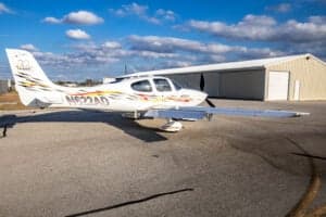 A Cirrus SR22 aircraft with a custom paint scheme is parked on the tarmac in front of a hangar.