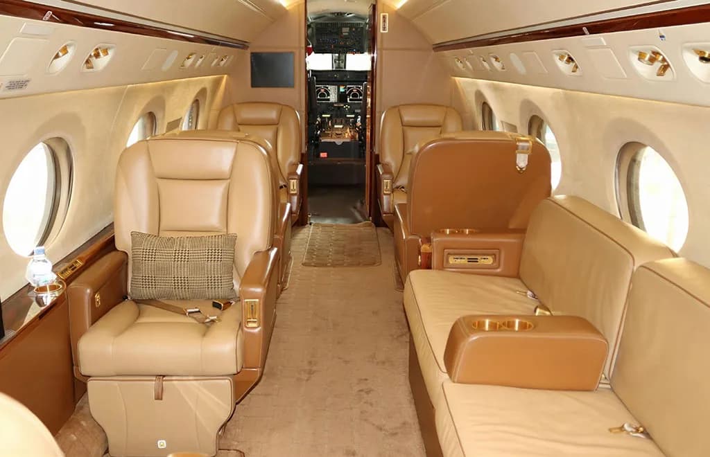 The spacious interior of a private jet, showing tan leather seating, a sofa, and a glimpse of the cockpit.