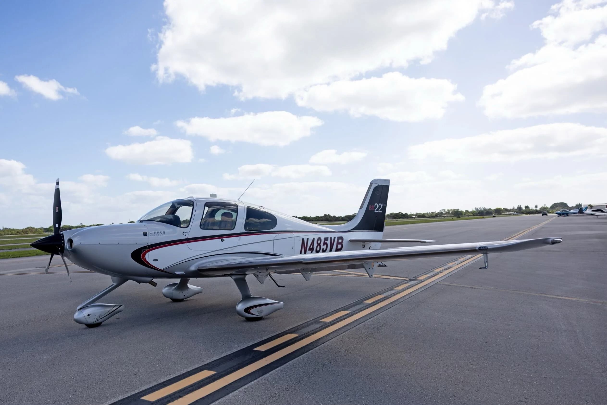 A side-quarter view of the silver Cirrus SR22 GTS aircraft N485VB, parked on an airfield under a bright, partly cloudy sky.
