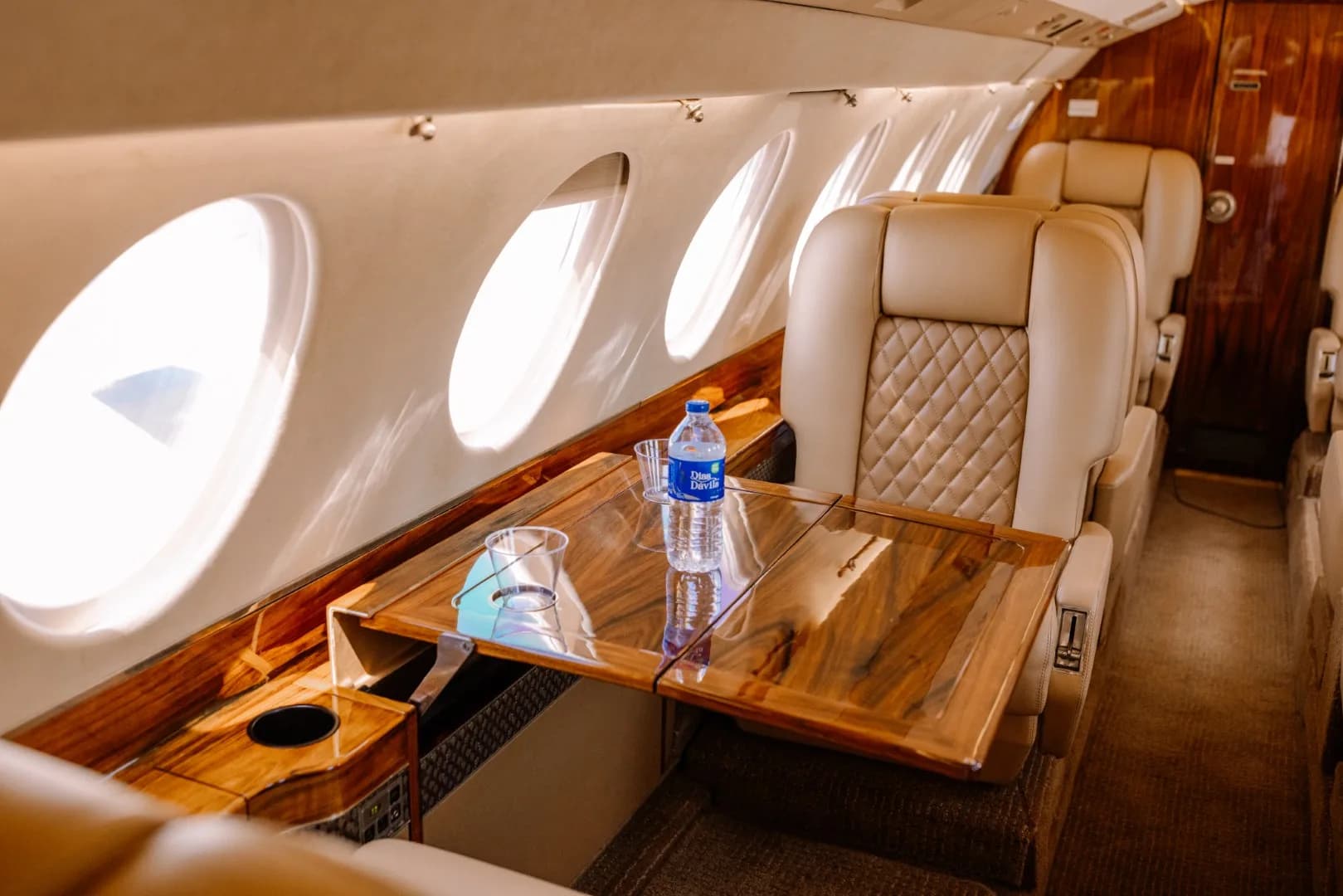 A polished wooden table extended from the side of an aircraft cabin, with a bottle of water and a glass.