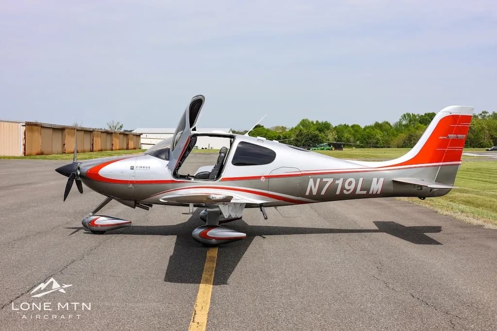 Side view of a silver and orange Cirrus SR22 aircraft with doors open, parked on an asphalt runway.