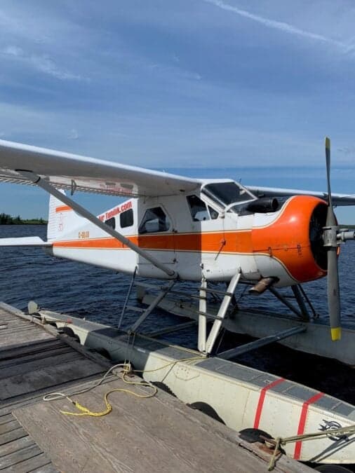 A de Havilland Beaver floatplane is docked on a wooden platform on a lake under a blue sky.