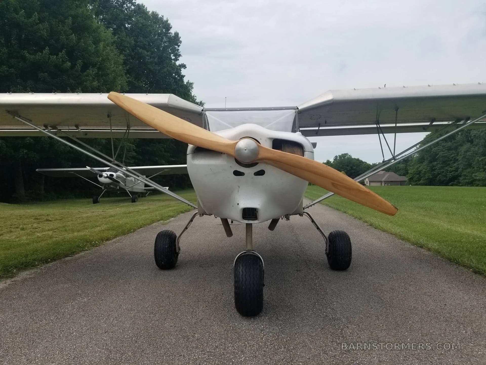 A head-on view of a light gray ultralight aircraft with a large wooden propeller, parked on a paved path with another aircraft in the background.