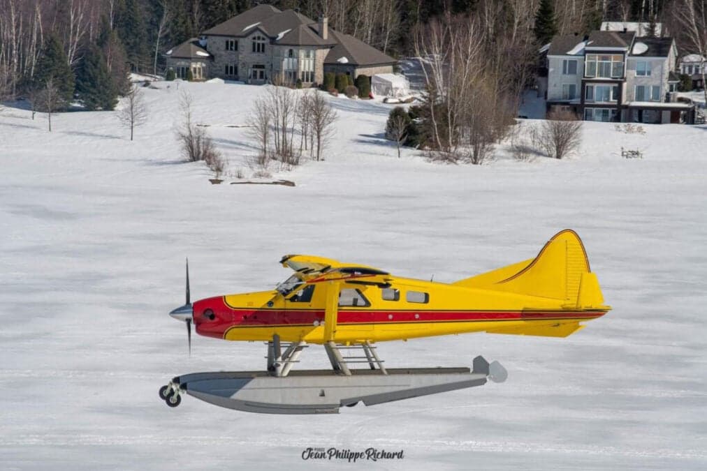 Yellow and red floatplane on a frozen lake with houses in the background.