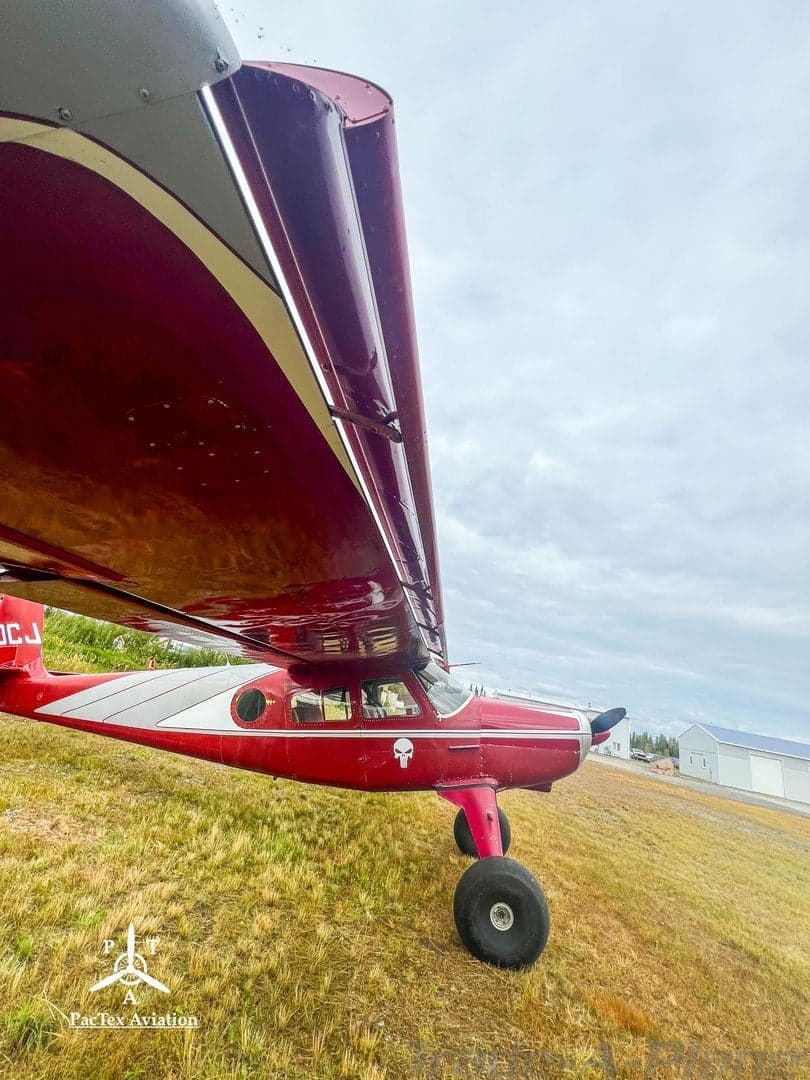 A low-angle view of the rear-left side of a red and silver single-engine aircraft, showing the underside of the wing and the tail.