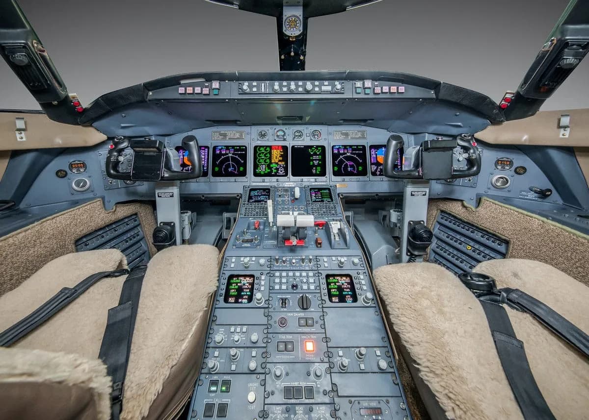 The cockpit of a business jet, showing multiple digital displays, control yokes, and a central control panel with numerous buttons and switches.