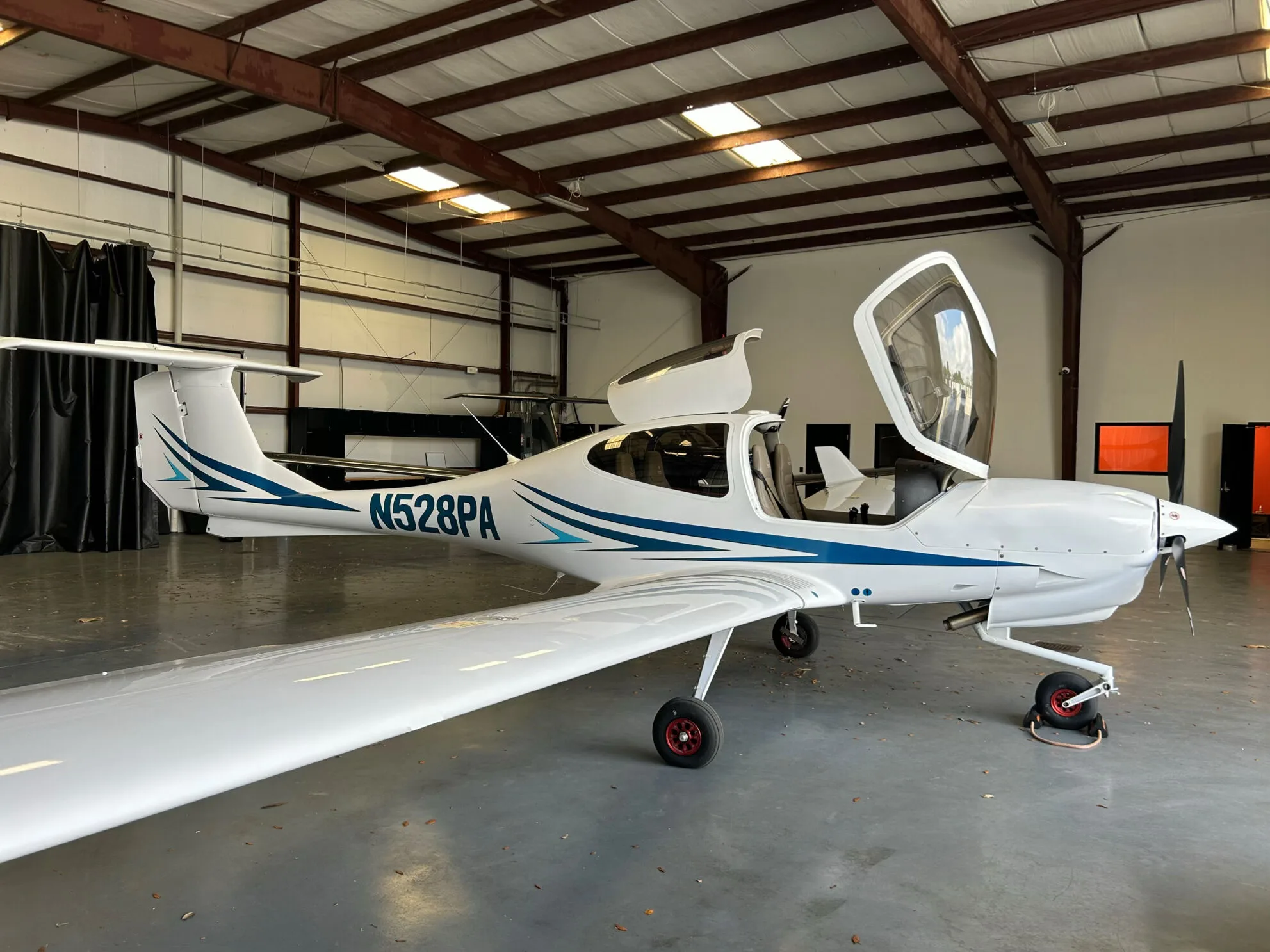 Side view of a white Diamond DA40 aircraft with blue and light blue stripes, registration N528PA, parked in a hangar with its canopy open.