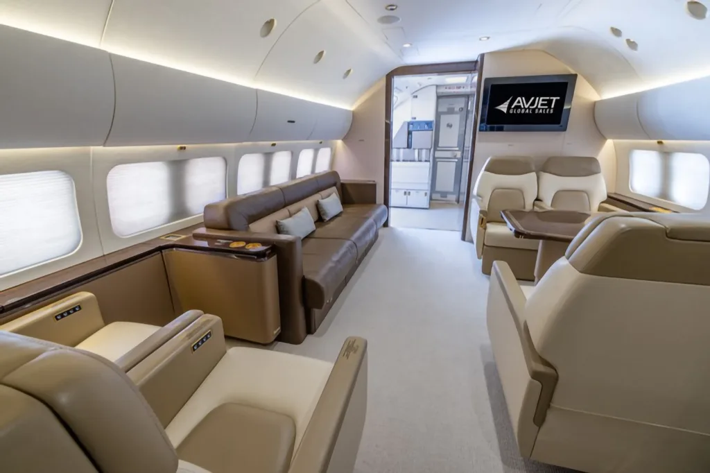 A spacious aircraft interior with a brown leather sofa, individual beige seats, and a screen displaying the Avjet Global Sales logo.