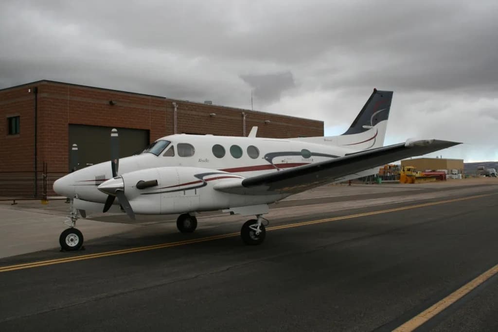 A side view of a white twin-engine turboprop aircraft with dark red and blue stripes, parked on an asphalt runway next to a brick building.