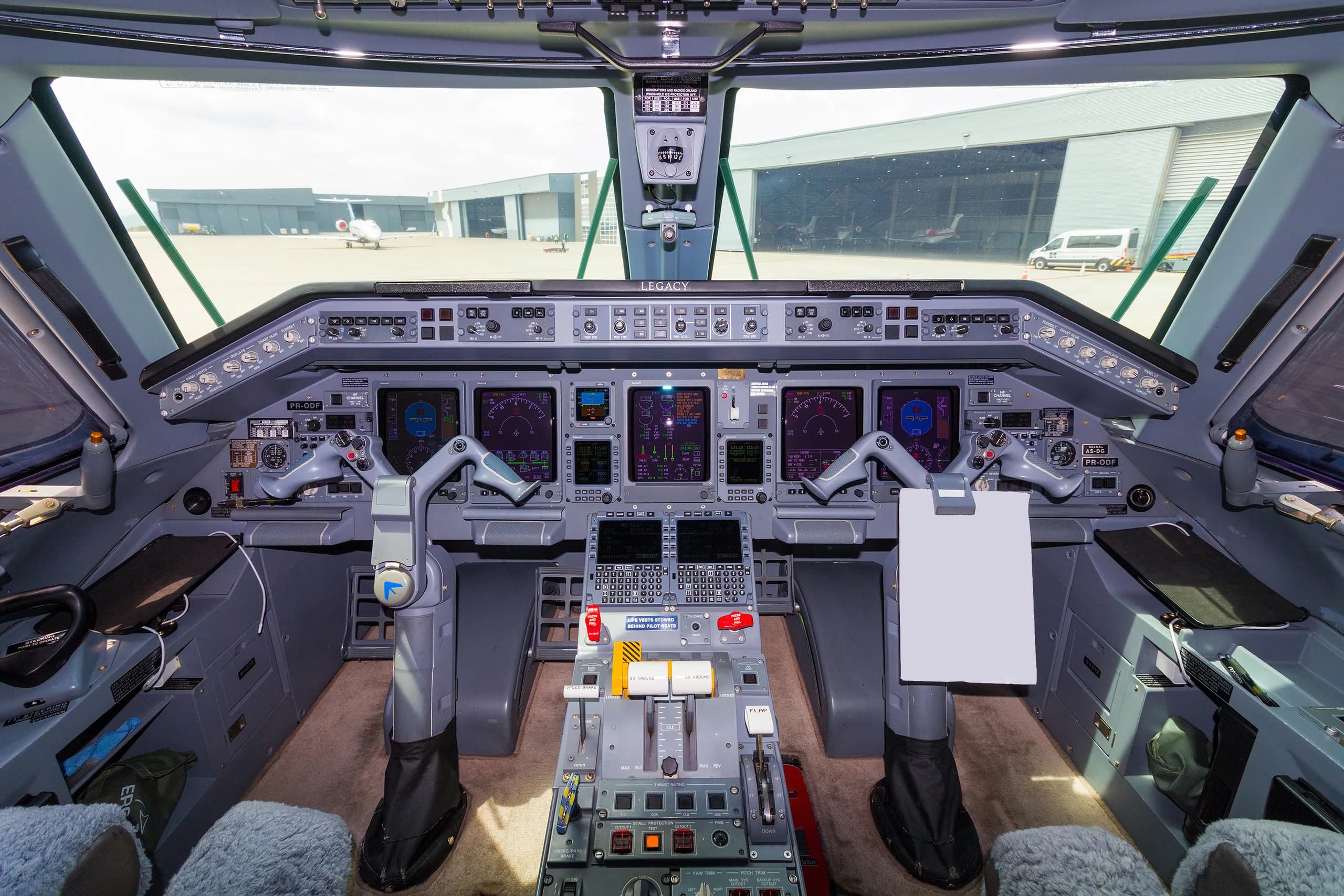 The cockpit of an Embraer Legacy aircraft, showing the instrument panel, yokes, and view of hangars outside.