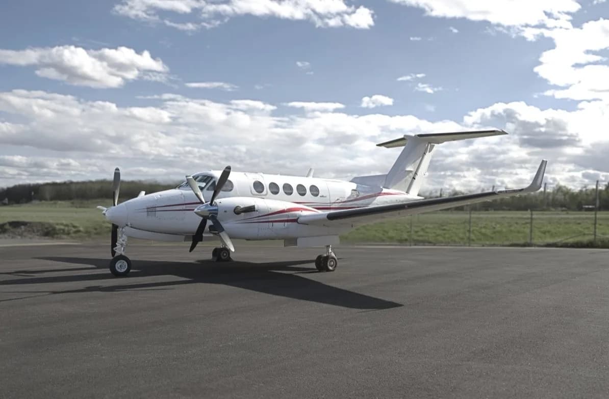 A white twin-turboprop aircraft with red accent stripes is parked on a tarmac under a partly cloudy sky.