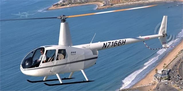 A white Robinson R44 helicopter flying over the ocean.