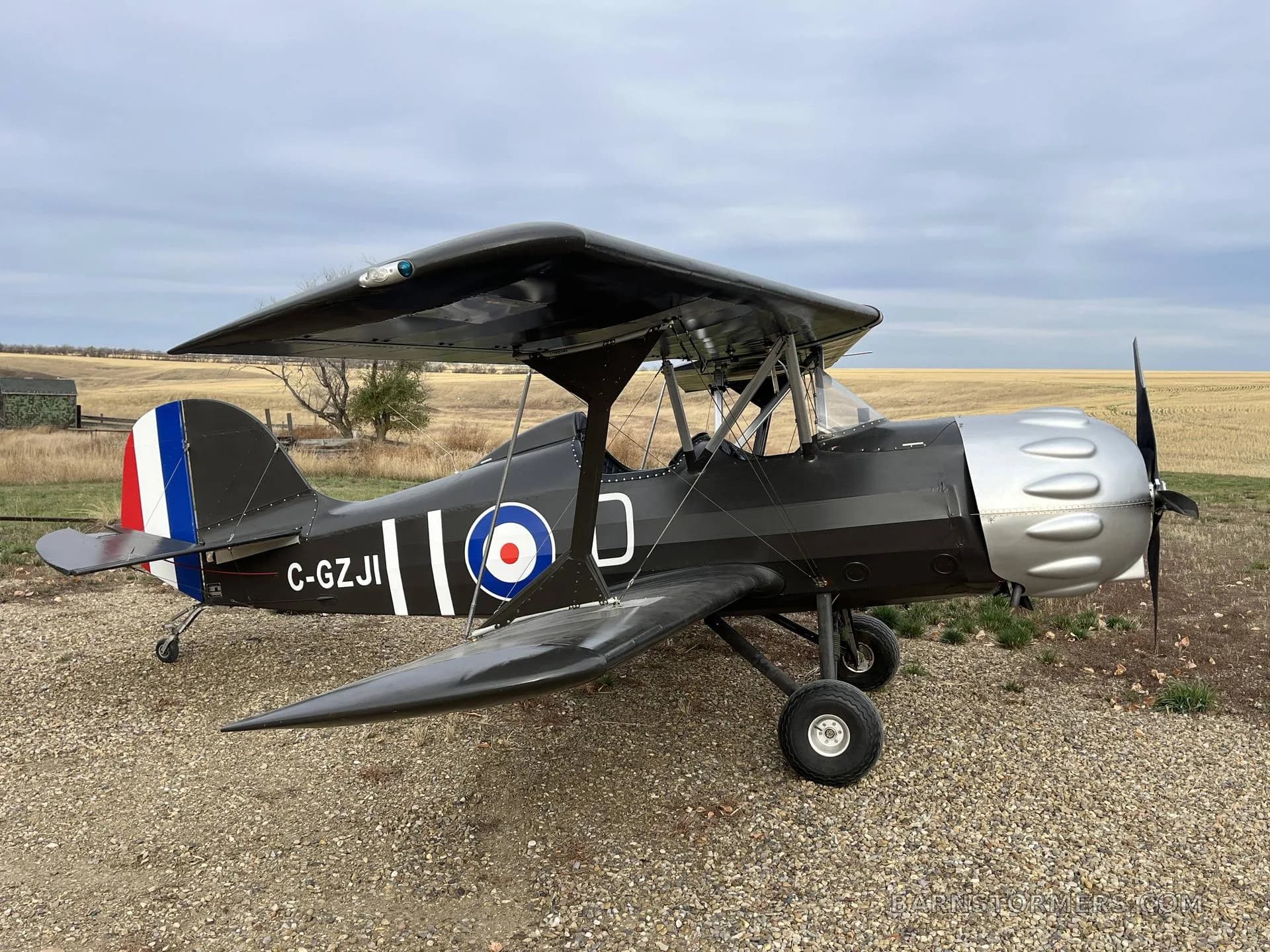 A black biplane with a silver engine cowling and British roundel markings, parked on a gravel path in a field under a cloudy sky.