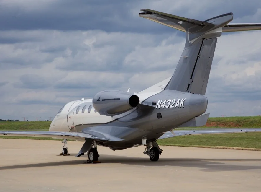 A rear three-quarter view of the white and grey Embraer Phenom 300E business jet, N432AK, on an airfield.