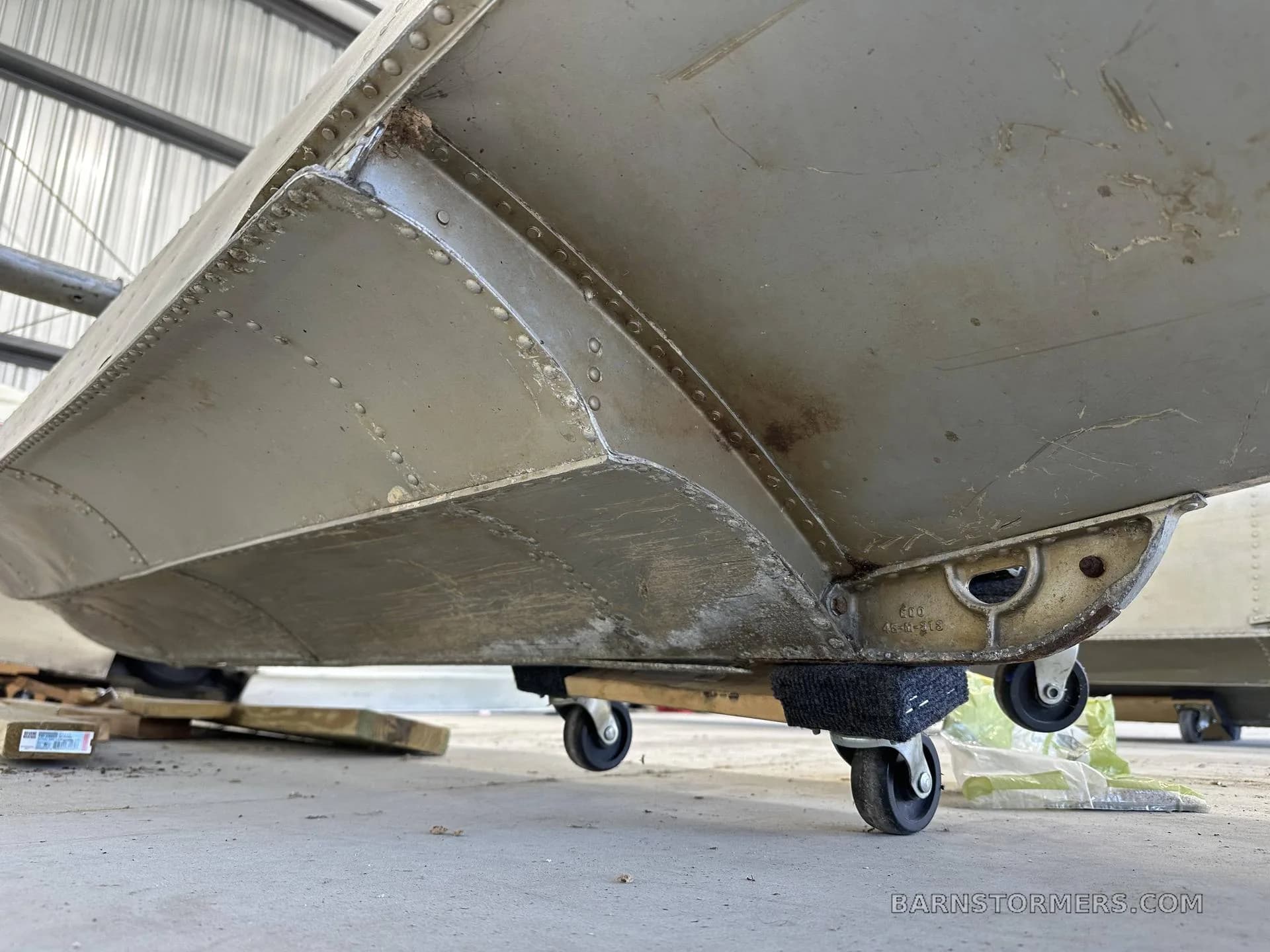 The underside of an aircraft float, showing its construction and a dolly with wheels, is in a hangar.