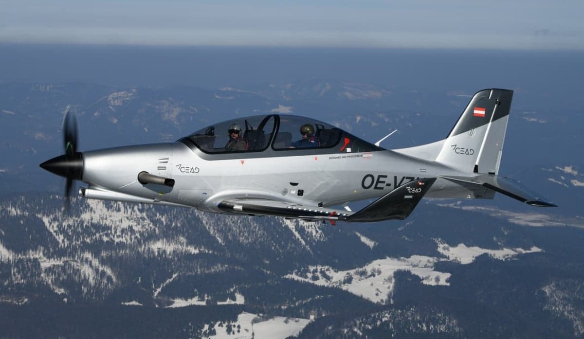 A silver CEAD aircraft flies over a snowy mountain range.