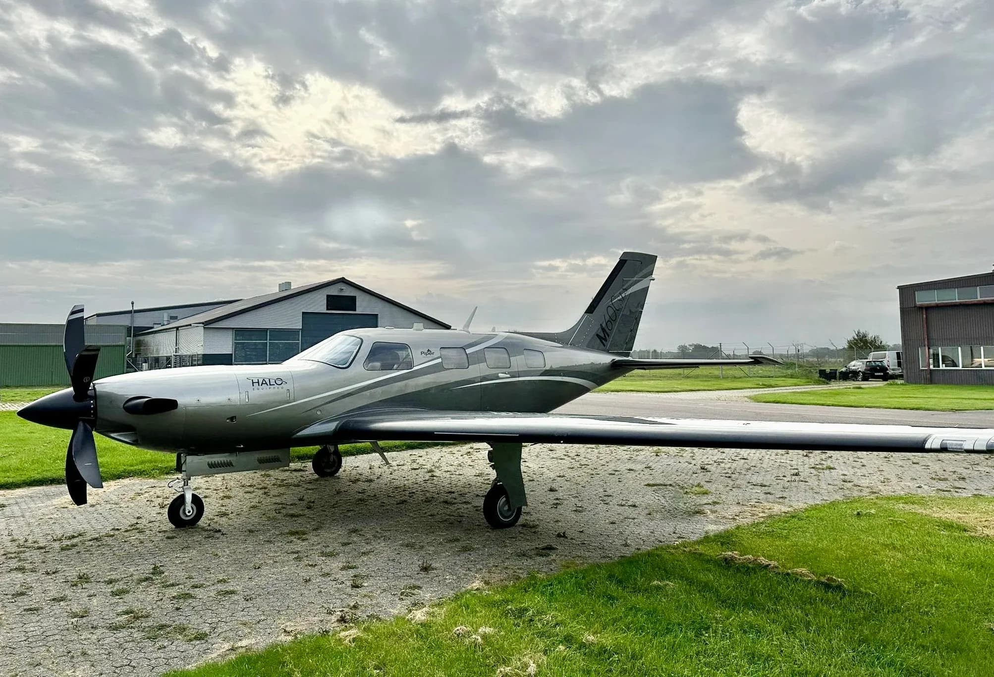 A Piper M600 HALO aircraft parked on the grass in front of a hangar on a cloudy day.