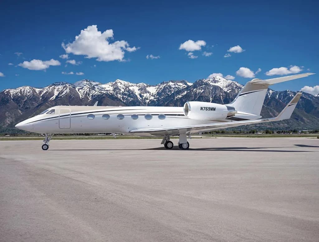A side view of a white Gulfstream G650 business jet with registration N769MM on a tarmac, with mountains under a blue sky.