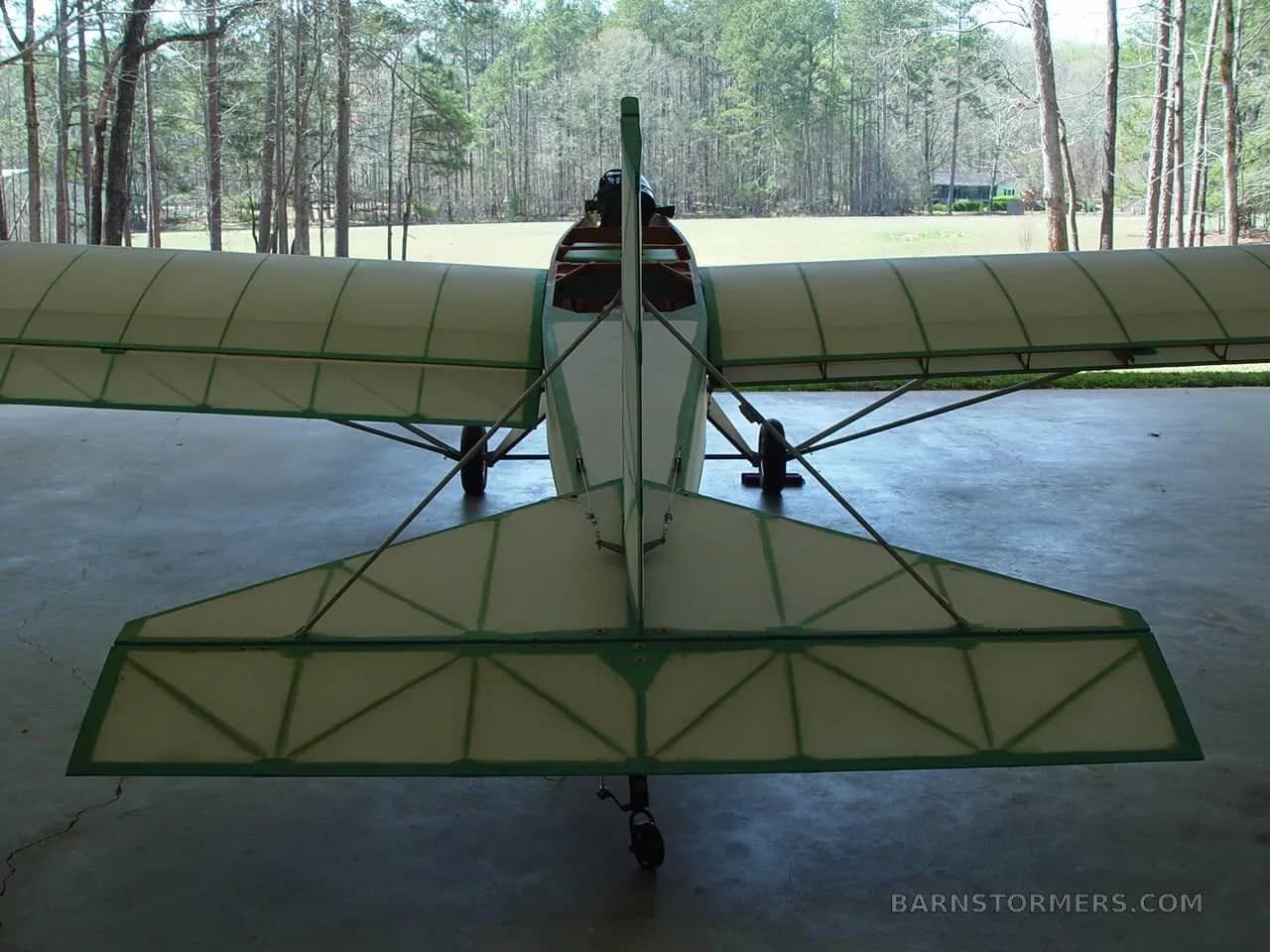 A rear view of a light aircraft with a wooden fuselage and green trim, parked in a hangar with trees in the background.
