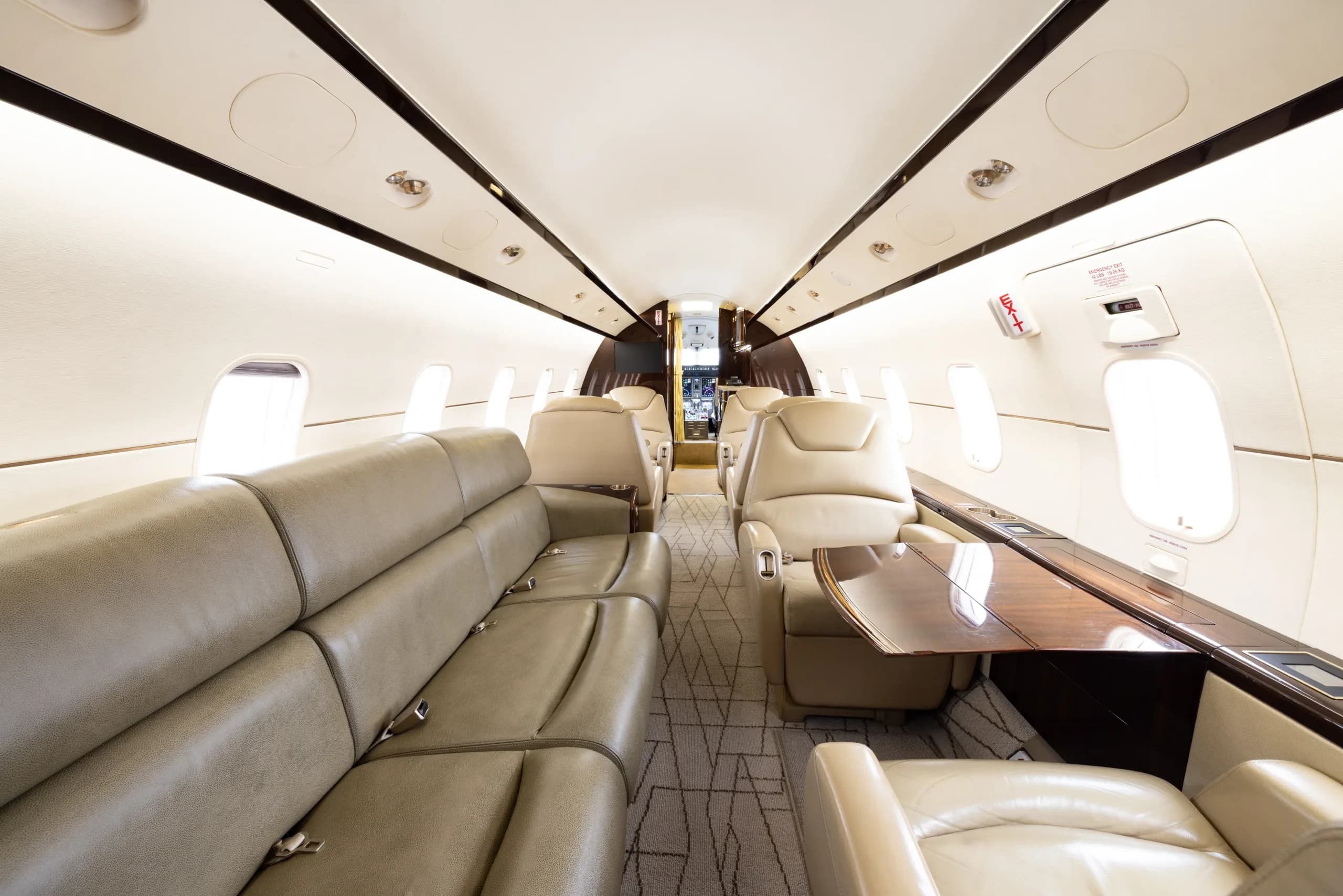 The spacious interior of a business jet cabin with a long beige leather sofa and individual seats with wooden tables.