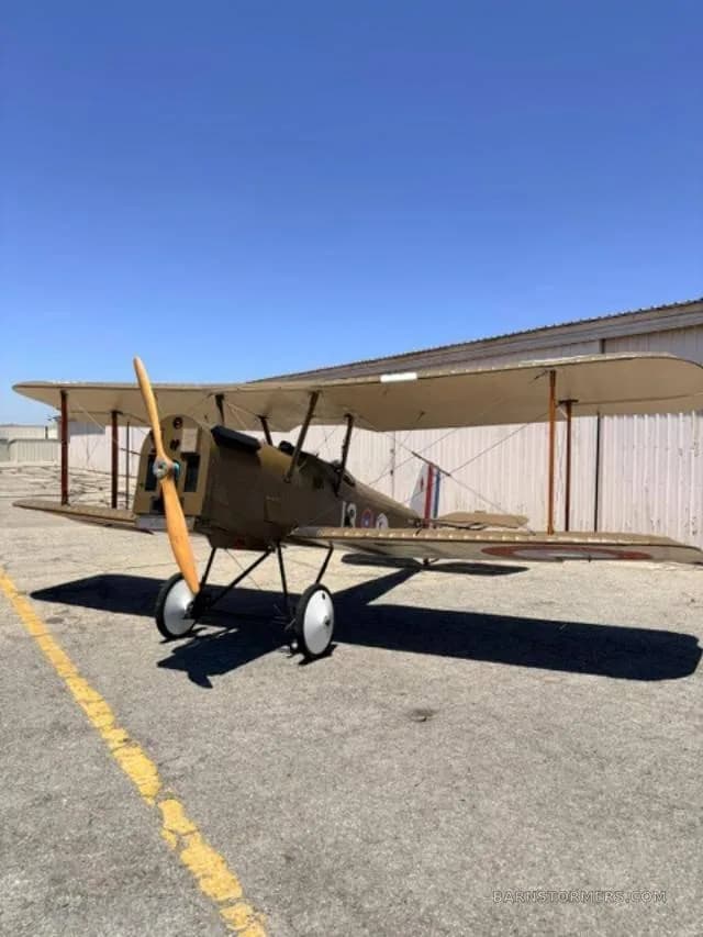 A front-side view of a brown biplane with a wooden propeller and white wheels, parked on an asphalt apron.