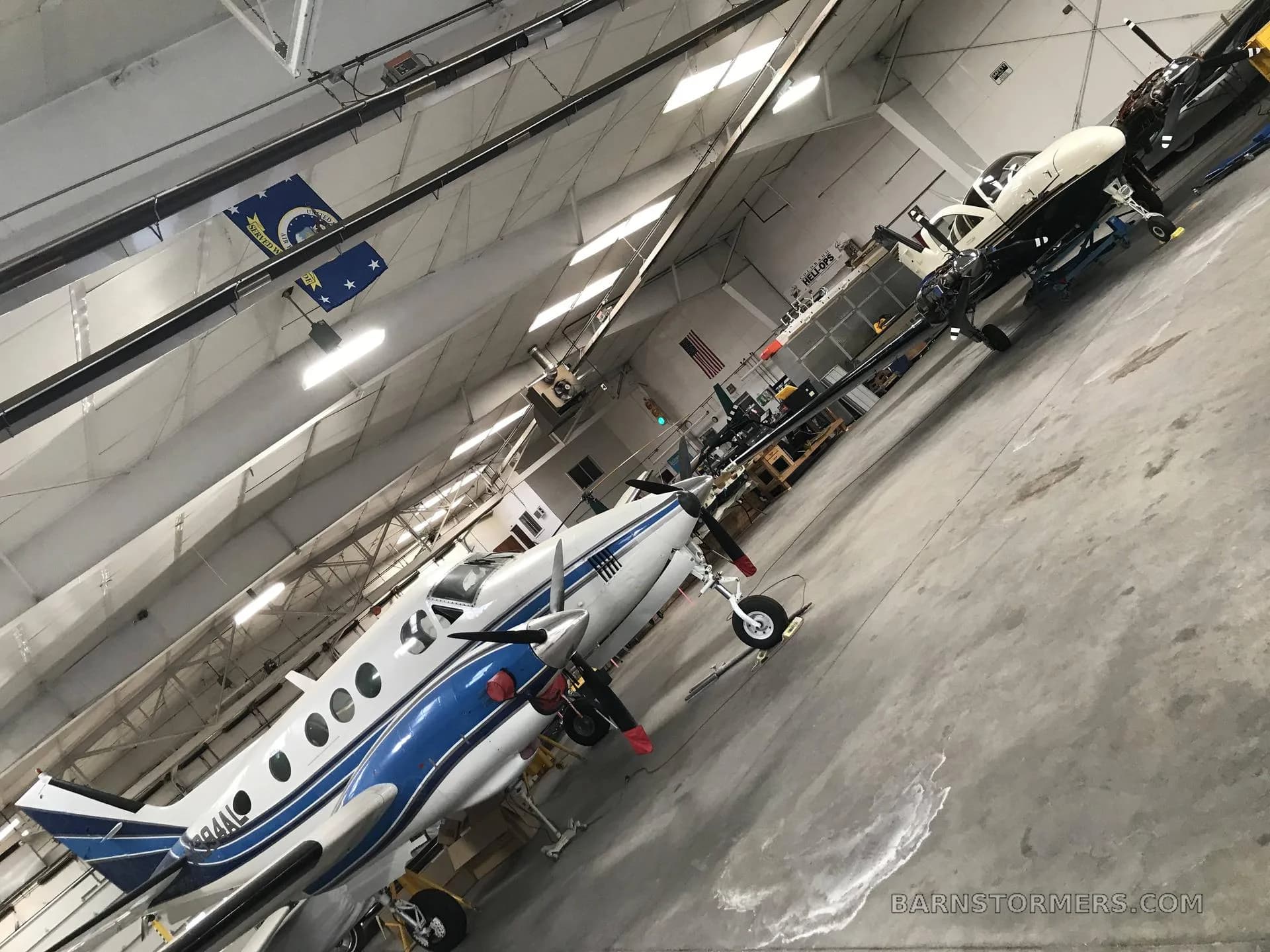 A white and blue twin-engine turboprop aircraft is parked in a hangar, with another aircraft visible in the background.