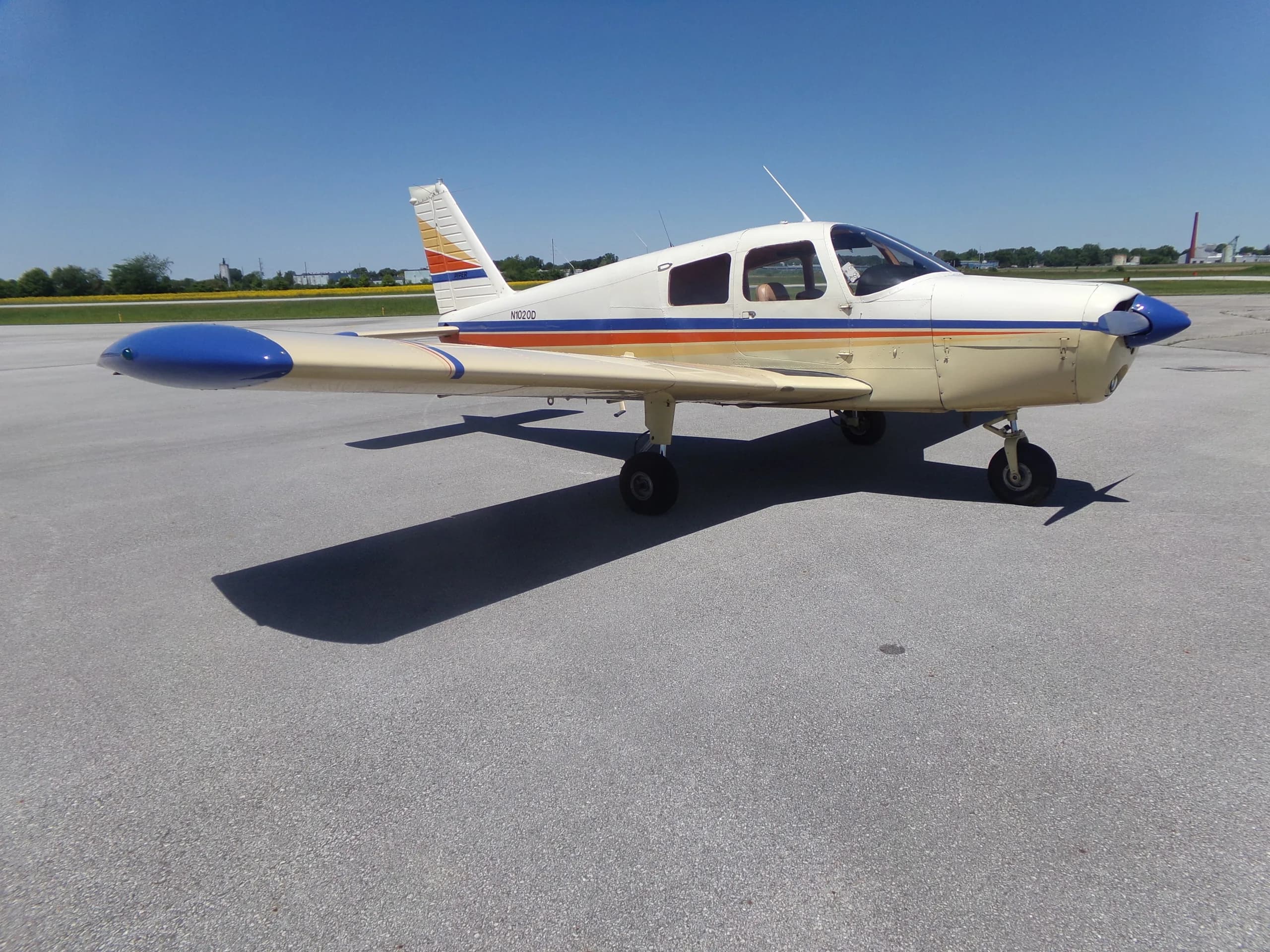 A white and beige Piper aircraft with blue, orange, and yellow stripes, seen from the front-right, parked on an asphalt tarmac.