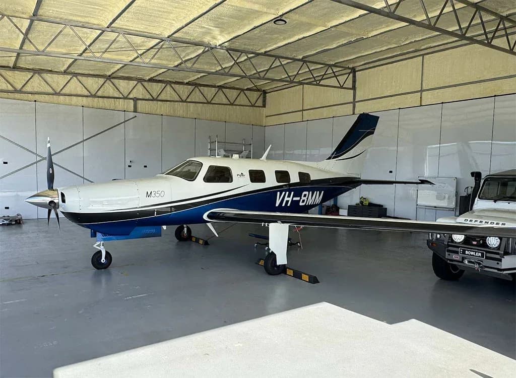 A white and blue Piper M350 aircraft, registration VH-8MM, is parked inside a hangar next to a white Land Rover Defender.