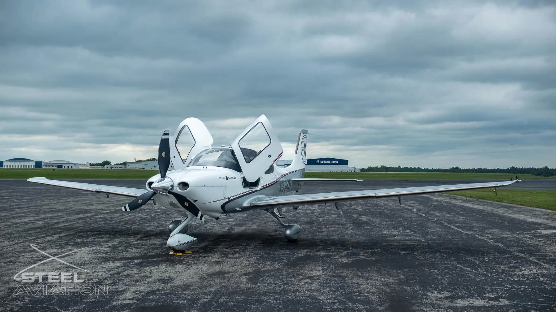 A white and silver Cirrus SR22T aircraft with its gull-wing doors open is parked on an airfield under a cloudy sky.