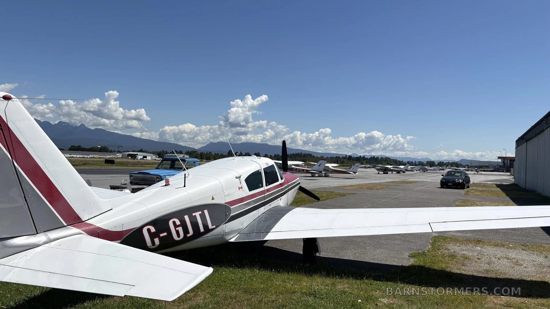 Rear view of a white and red single-engine propeller aircraft (C-GJTL) parked on an airfield, with other aircraft and mountains in the distance.
