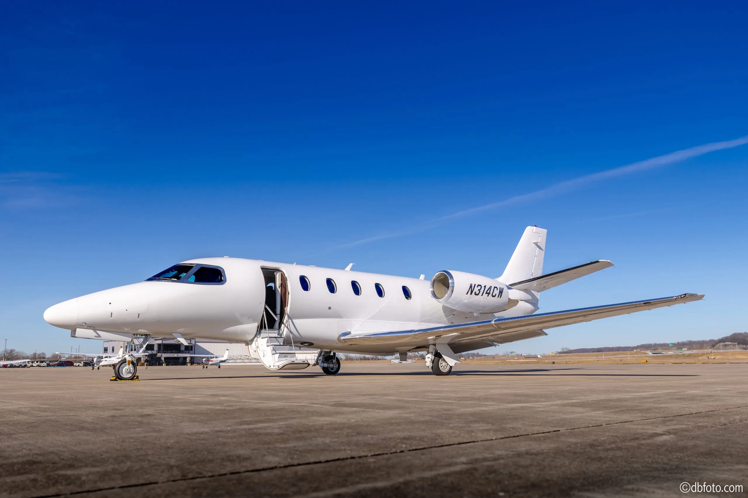 A sleek all-white Cessna Citation business jet with its passenger door open, parked on an airfield under a clear blue sky.