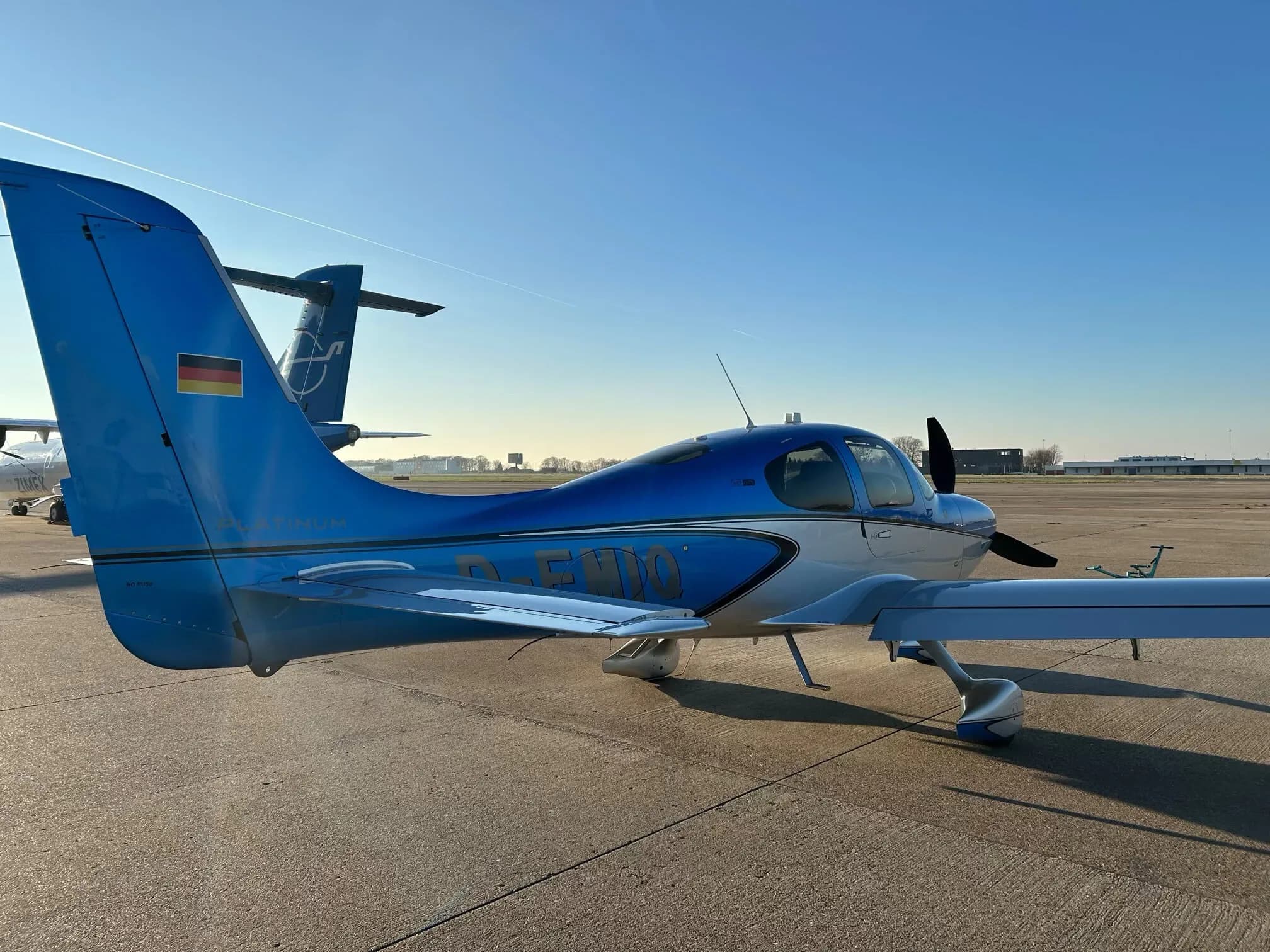 A rear-side view of a blue and silver Cirrus SR22 aircraft with the German flag on its tail, parked on an airfield.