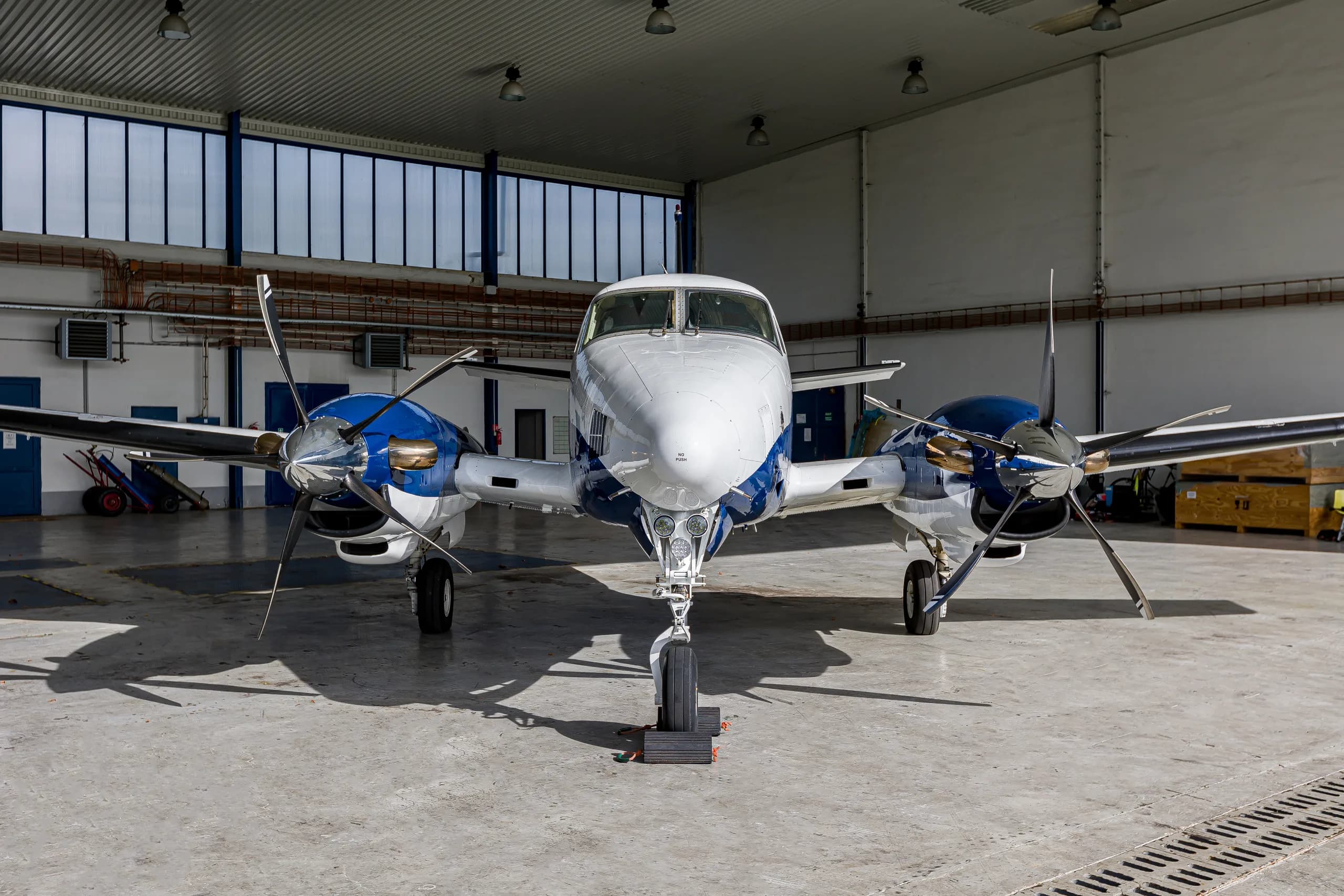 A head-on view of a white and blue twin-engine turboprop aircraft, likely a King Air, parked inside a hangar.
