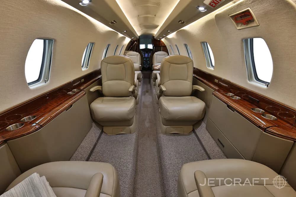The aircraft cabin features beige leather seats, wood trim, and overhead lighting, looking towards the cockpit.