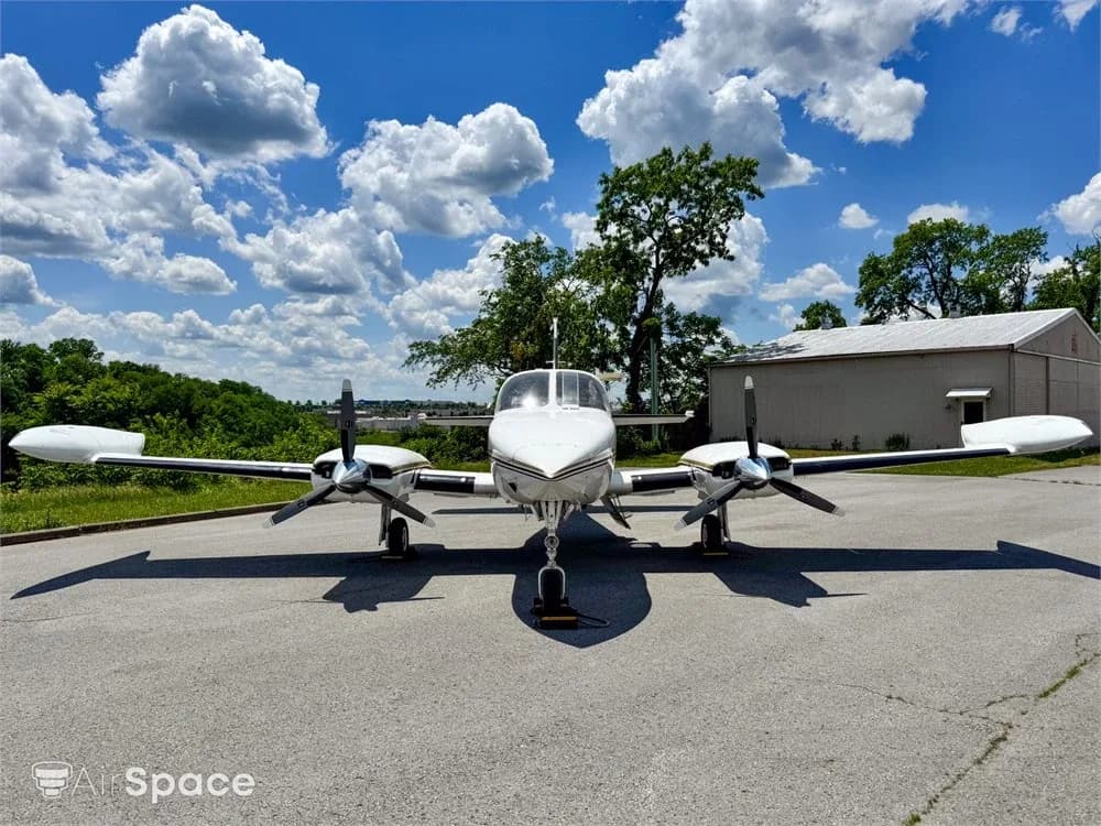 A front view of a white twin-engine aircraft with gold and brown stripes parked on an asphalt ramp under a partly cloudy sky.