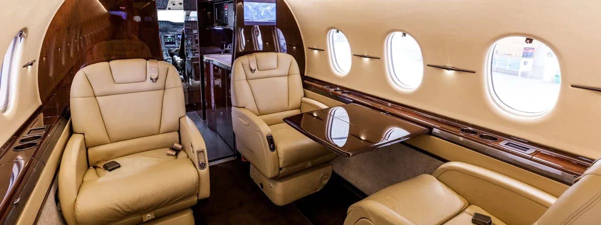 A wide shot of an aircraft's luxurious cabin interior featuring tan leather seats, a polished wood table, and a view towards the cockpit.